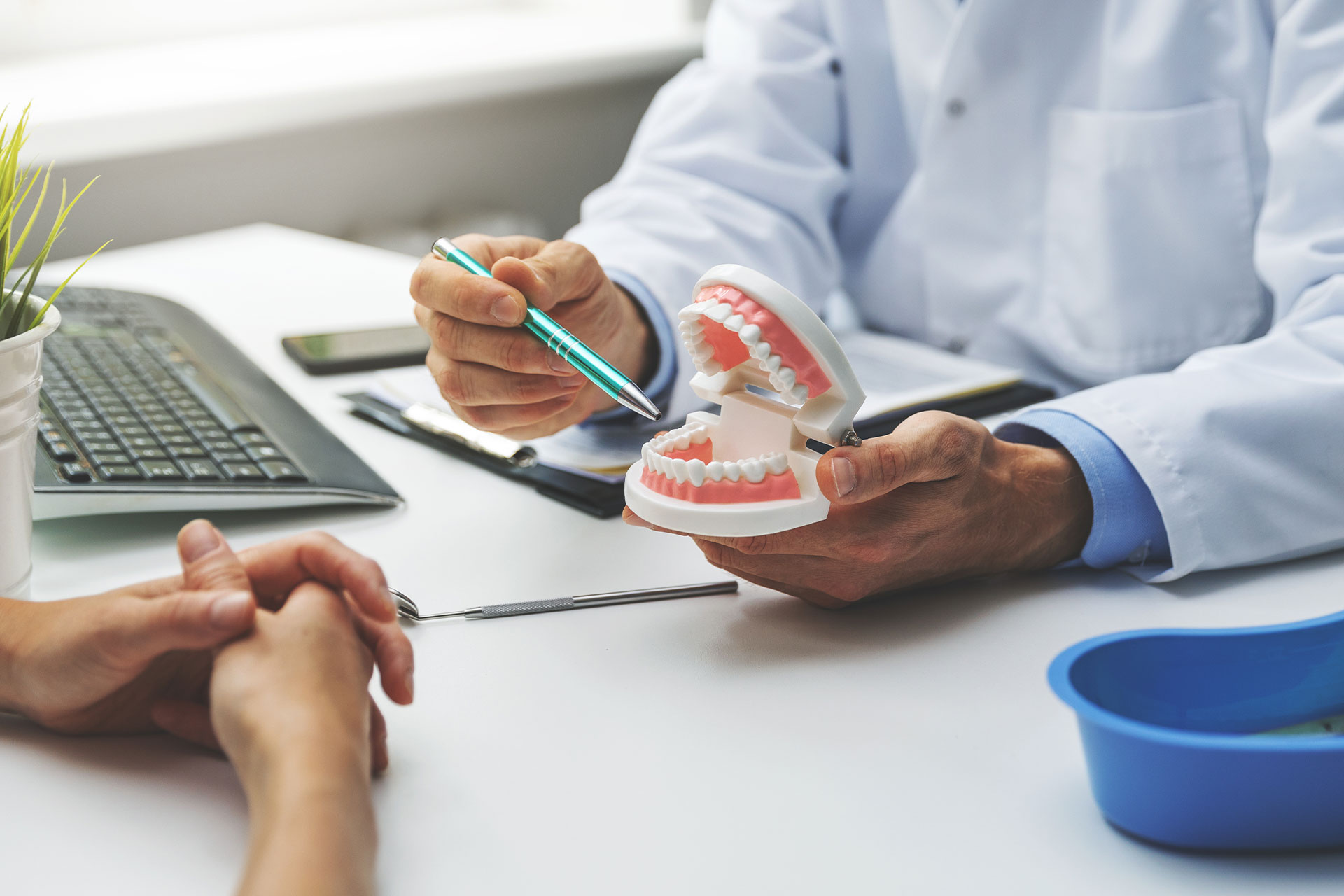 A dental professional in a white coat and stethoscope, seated at a desk with a model of human teeth, holding a dental tool, while another person s hands are visible, suggesting an interaction or examination.