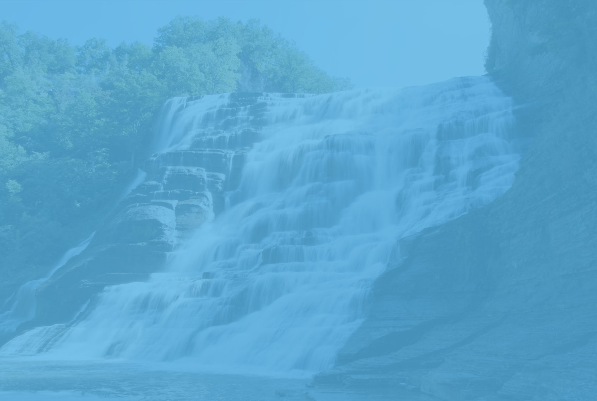 A waterfall cascading down a cliff, surrounded by lush greenery and a clear blue sky, with the bottom of the image showing a portion of a river.