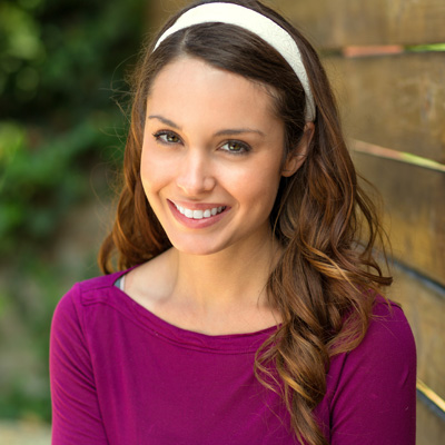 A woman with long hair, wearing a purple top and headband, posing for a photo against a wooden fence.