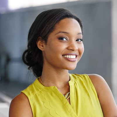 A young woman with a bright smile, wearing a yellow top and standing against a blurred background.
