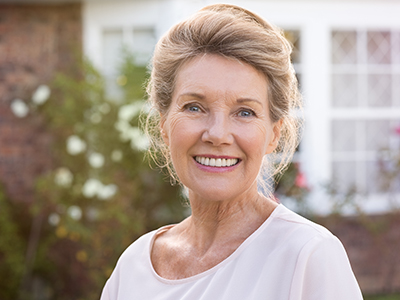 A woman with short hair, wearing a white top, stands in front of a brick house with a garden.