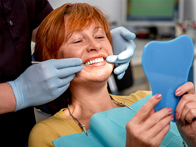 An elderly woman is seated in a dental chair, receiving oral care from a dentist who is adjusting her teeth with a mirror and blue tray.