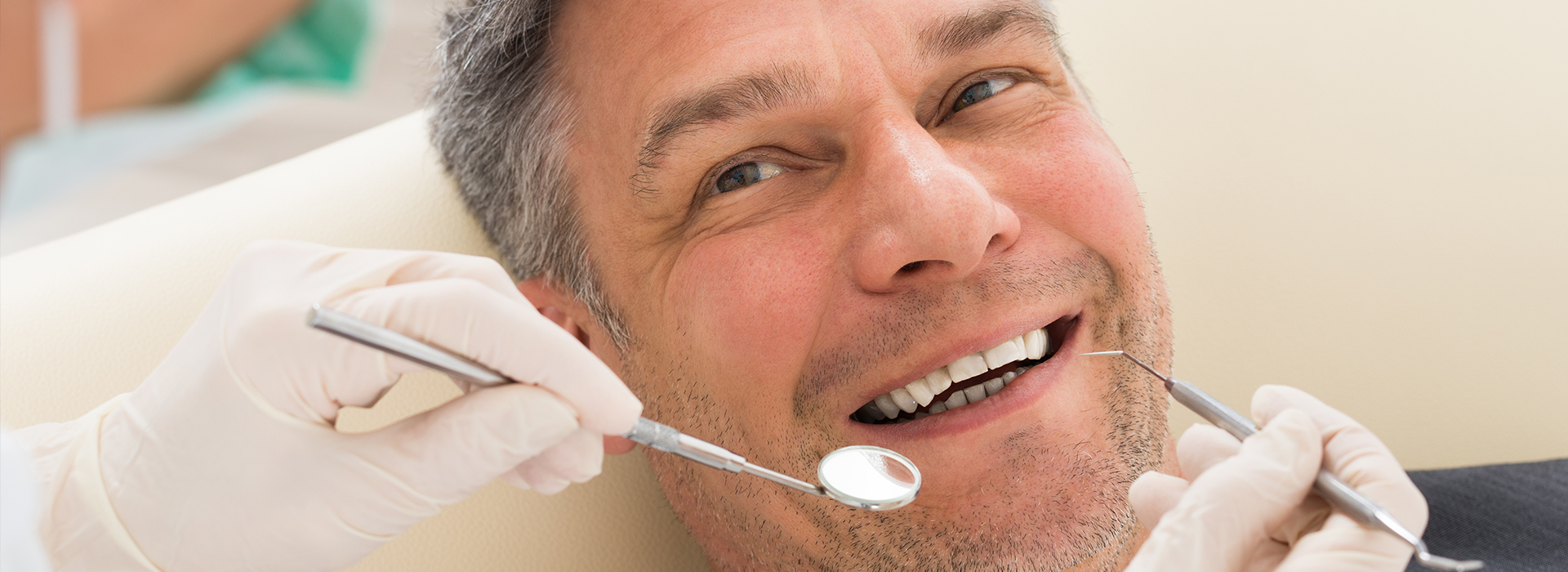 The image shows a man sitting in a dental chair, receiving dental treatment, with a smiling expression while wearing protective gloves and face mask.