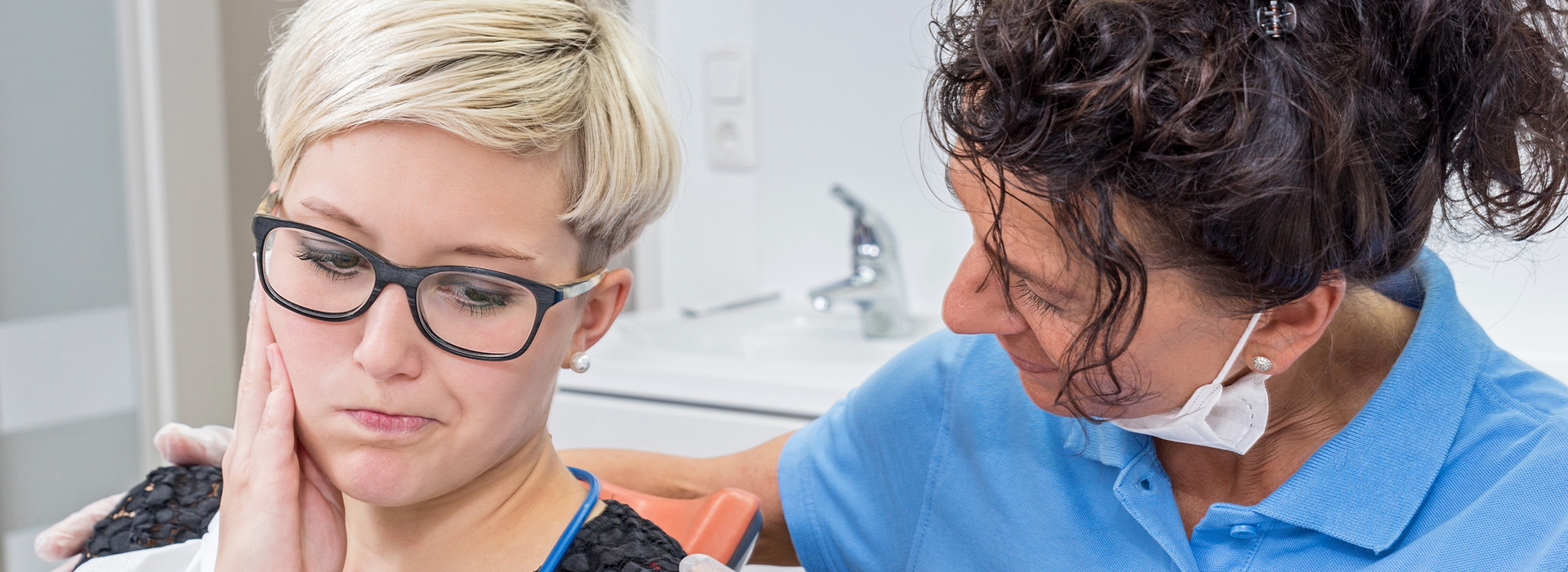 The image shows a woman with glasses sitting in a dental chair, receiving dental care from a female dentist who is performing a procedure on her teeth.