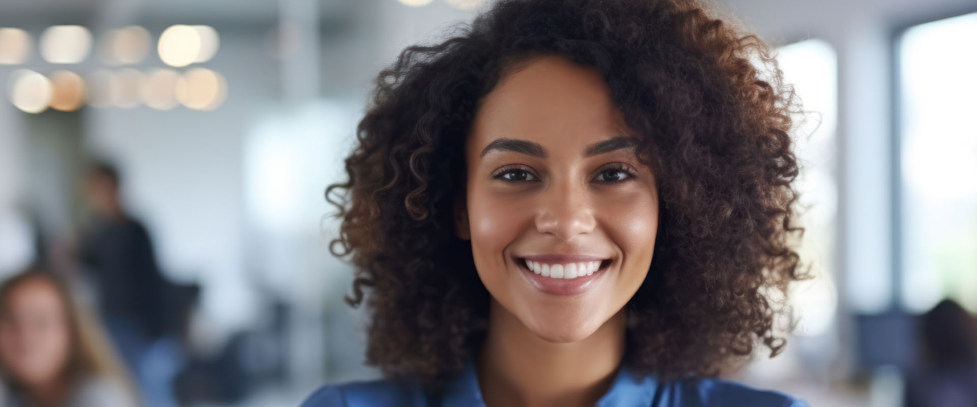 The image is a photograph of a smiling woman with curly hair, wearing a blue top and standing in an indoor office environment.