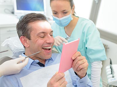 The image shows a man sitting in an oral care chair, holding up a pink card with a smile on his face, surrounded by dental professionals who appear to be engaged in a discussion or presentation.