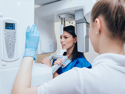 An image featuring a woman in a blue lab coat standing next to a large, modern 3D scanner machine, with another individual wearing gloves and a face mask observing the equipment.