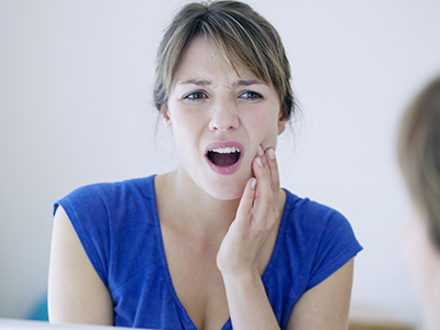 A woman in a blue top with her mouth open, showing teeth, and holding her hand to her face as if she s experiencing pain or discomfort.