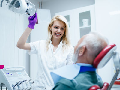 Woman in white scrubs assisting elderly man with dental procedure, both in dental office.