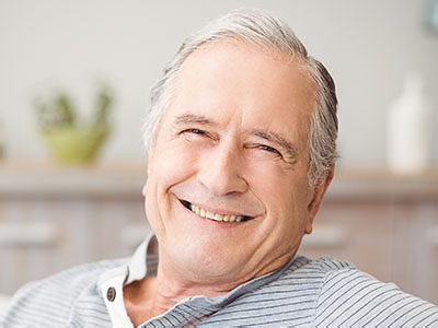 The image shows an elderly man with a smile, seated comfortably in a chair and looking directly at the camera.